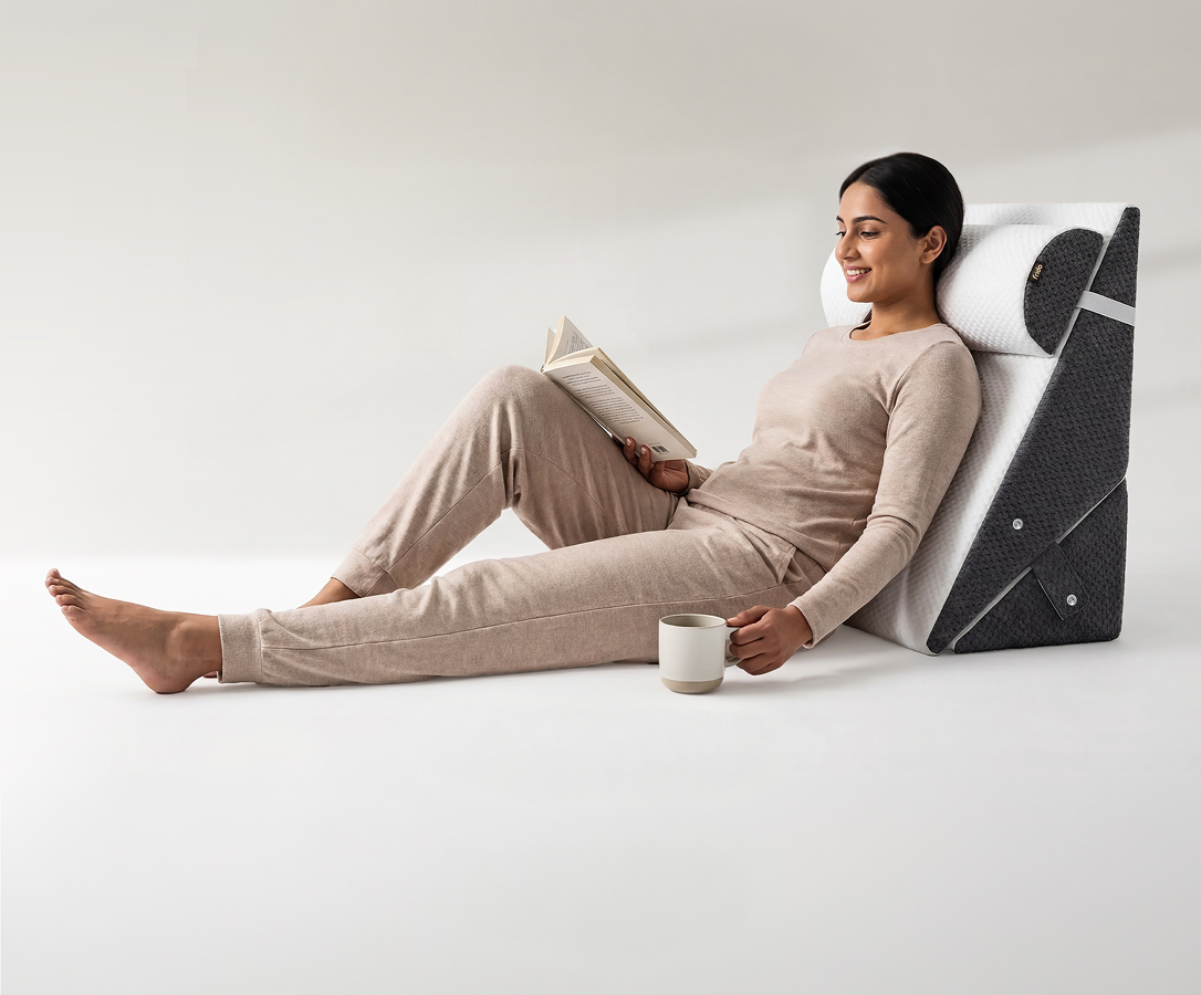 A woman in a beige pajama set reclines against a white and black wedge pillow while reading a book and holding a mug in a bright, minimalist room.